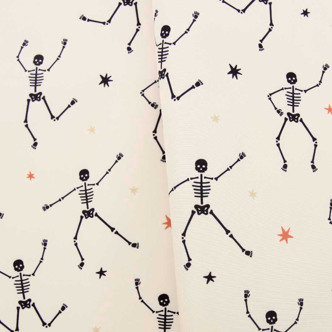 A close-up of a cream-colored fabric with a repeating pattern of black dancing skeletons and small black and orange stars.
